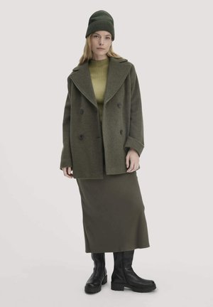 Doppelreihiger Mantel in Olivgrün mit großen Revers, kombiniert mit einem hellolivgrünen gerippten Pullover, einem langen olivgrünen Rock und schwarzen Stiefeln.
