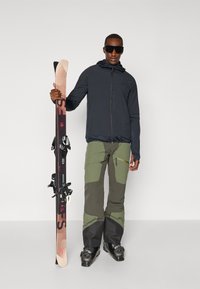 Slidinimo apranga, kurią sudaro tamsiai mėlynas zip-up striukė, žali ir juodi slidinėjimo kelnes su sustiprintomis dalimis, ir slidinėjimo batai. Laiko slidės su tvirtinimais.