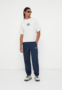 T-shirt grigia a maniche corte con logo blu navy, abbinata a pantaloni da jogging blu navy con logo bianco. Sneakers grigie e bianche completano l'outfit.