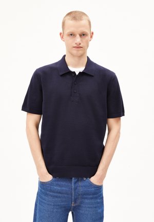 Marineblaue Kurzarm-Polohemden mit klassischem Kragen, einer Drei-Button-Leiste und geripptem Saum. Getragen mit blauen Jeans.