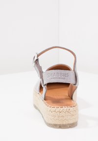 Šedá semišová sandál s texturovanou espadrilovou podrážkou a nastavitelným pásem na kotníku, obsahující koženou stélku a logo na pásku.