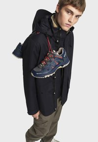 Mornarica vodootporna jakna s kapuljačom, bež unutrašnjost i sivi planinarski cipele s crvenim vezicama. Cipele imaju teksturiranu potplatu i mrežaste panele.