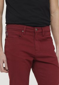 Jean en coton rouge avec une surface texturée, conception à cinq poches, fermeture à bouton en métal et étiquette logo brodée sur la poche avant.