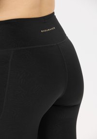 Close-up van een persoon die hoge zwarte leggings draagt met subtiele patronen en het woord "ENDURANCE" op de tailleband.