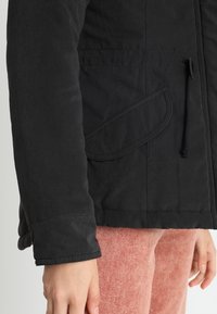 Manche de veste noire à rabat sur pantalon rose texturé, montrant une partie du bras et de la main détendue sur le côté.