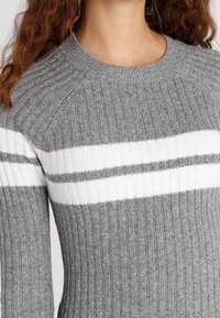 Grå stickad tröja med vita och ljusgrå horisontella ränder. Har ribbat mönster och rundad halsringning.