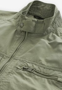Veste vert olive dotée d'un col haut, d'une fermeture éclair frontale et d'une poche latérale avec fermeture éclair. Fabriquée en tissu léger et texturé.