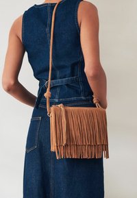 Borsa a tracolla in suede marrone con dettagli in frange, forma rettangolare e chiusura con zip. Indossata con un outfit in denim.