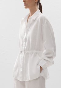 Chemise en lin blanc avec un devant boutonné, des manches longues et une taille à cordon. Elle présente une coupe décontractée et un tissu texturé.