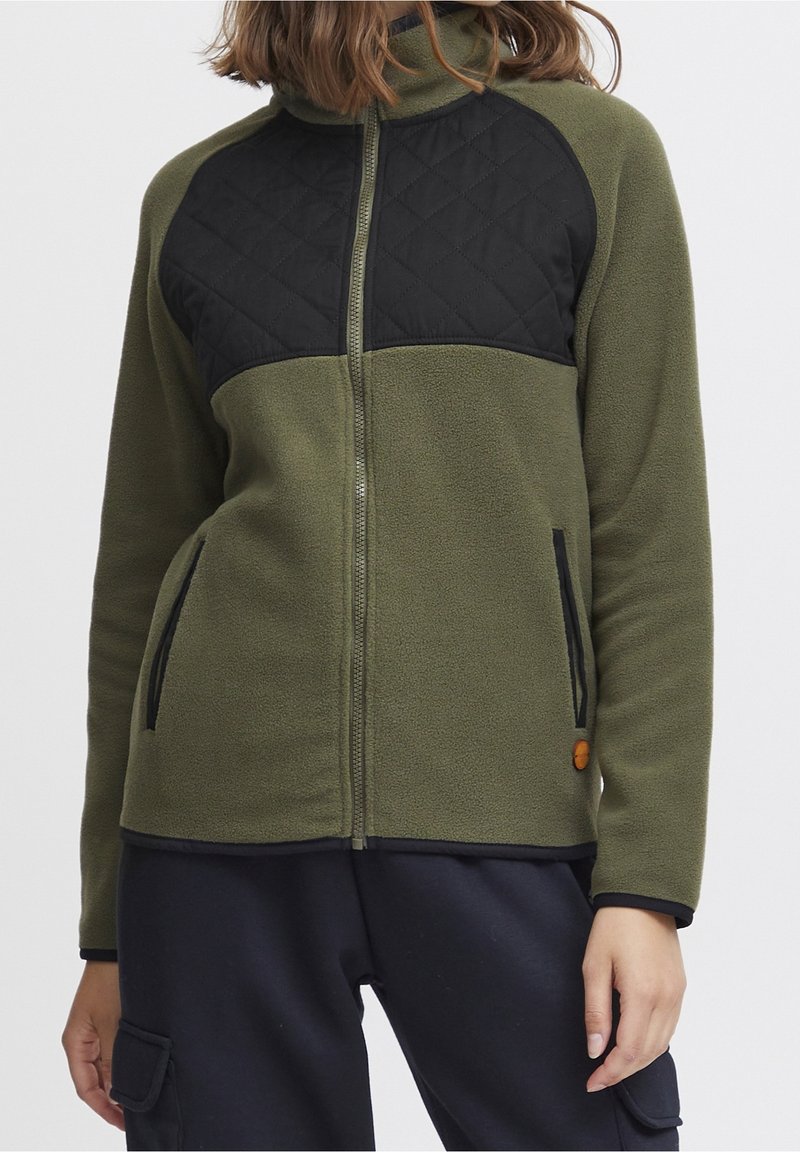 Femme portant une veste polaire vert olive avec un panneau matelassé noir sur la poitrine et des poches zippées, associée à un pantalon noir.