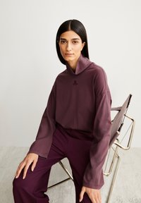 Übergroßes, langärmliges Oberteil aus tiefpurpurfarbenem Stoff mit hohem Kragen und dezent gesticktem Logo. Kombination mit passenden lila Hosen.