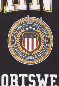 Emblema circular con escudo que lleva "1949", estrellas y una corona de laurel, rodeado de texto: "En el espíritu de la moda deportiva americana."