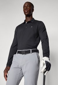 Μαύρο πόλο μπλουζάκι με μακριά μανίκια και μικρό λογότυπο, συνδυασμένο με ανοιχτό γκρι παντελόνι και μαύρη ζώνη. Λευκό γάντι γκολφ σε ένα χέρι, κρατώντας μια ρακέτα.