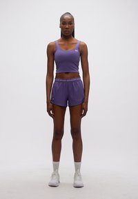 Γυναίκα που στέκεται με το πρόσωπο προς τα εμπρός, φοράει μοβ αθλητικό τοπάκι και σορτς με λευκά αθλητικά παπούτσια σε απλό φόντο.