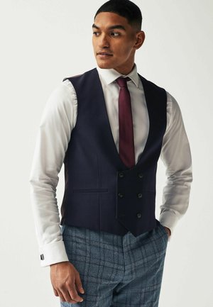 Navy vest with a deep V-neck, double-breasted buttons, and two front pockets. Worn over a white shirt and paired with gray plaid trousers.