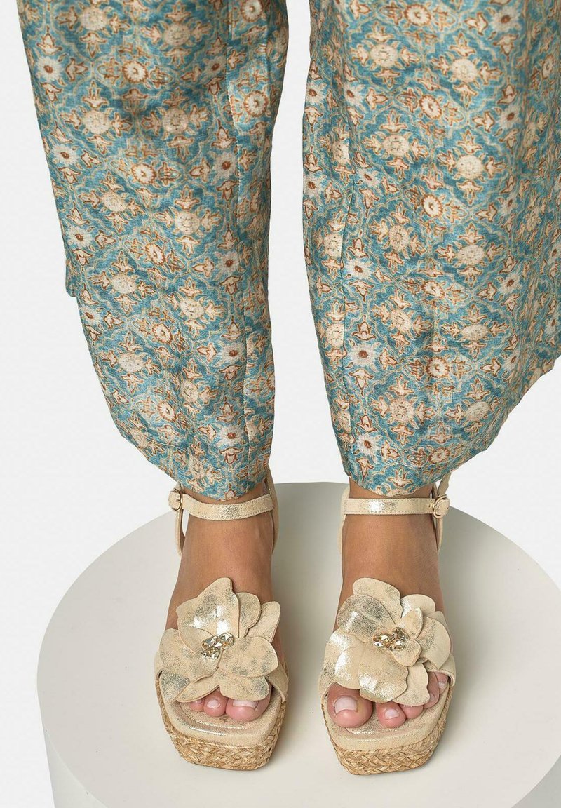 Pieds chaussés de sandales compensées beiges ornées de grandes décorations florales, associées à un pantalon ample bleu à motifs, coupé au-dessus des chevilles.