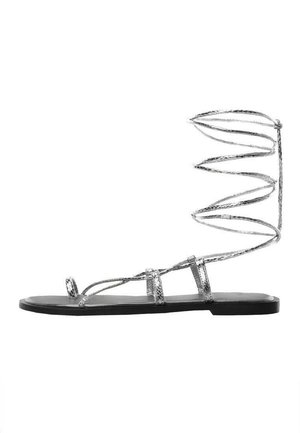 Silberne flache Sandale mit Riemchen und schwarzem Sohlen, ausgestattet mit einem gewundenen Knöchelriemen und mehreren dünnen Riemen über den Zehen und im Mittelfuß.