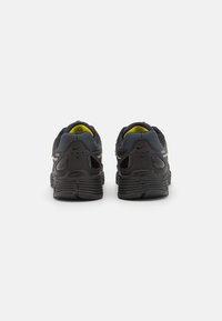 Black athletic shoes with a textured mesh finish, featuring a yellow accent and distinctive midsole design. Back view shows heel support details.