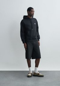 Μαύρη φούτερ με κουκούλα και μπροστινές τσέπες, συνδυασμένη με σορτς έως το γόνατο σε σκούρο χρώμα και δίχρωμα παπούτσια με λευκές κάλτσες.