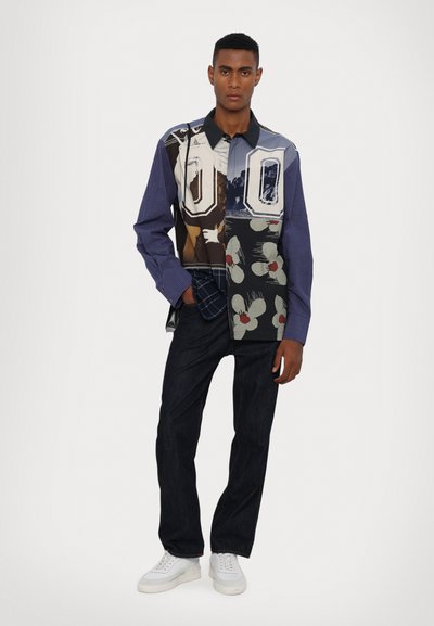 Multicolored button-up shirt featuring graphic prints, checkered patterns, and a dark blue sleeve. Paired with dark jeans and white sneakers.