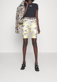 Černé priléhavé tričko, vzorované žluté a bílé cyklistické šortky s logy, černé sandály na podpatku a vzorovaná bunda přehozená přes jeden paži.