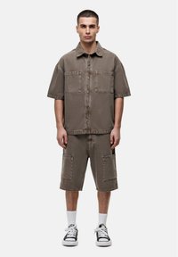 Bruine denim overhemd met korte mouwen en twee voorzakken, gecombineerd met bijpassende knielange shorts met grote zijzakken. Witte sokken en sneakers.