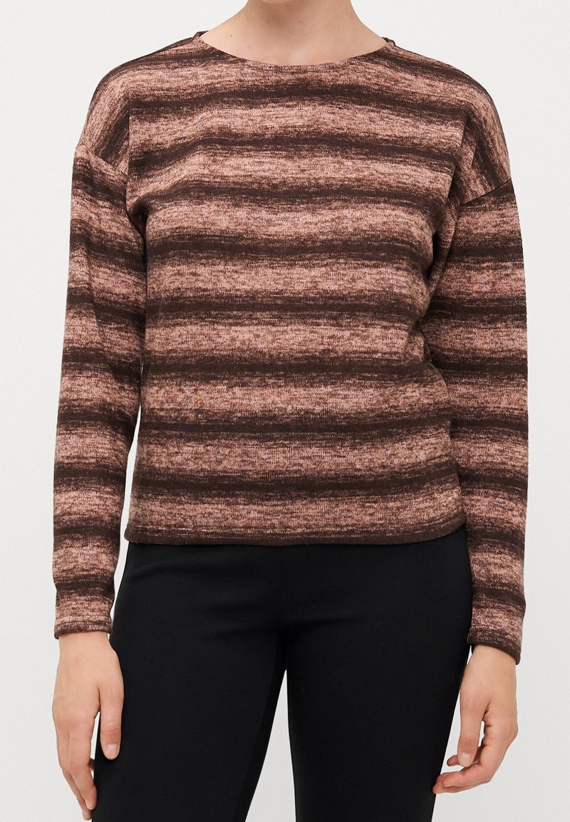 Femme portant un haut à manches longues avec des rayures horizontales marron et beige, associée à un pantalon noir, debout devant un fond uni.