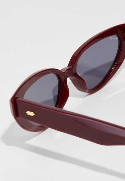 Lunettes de soleil en plastique rouge de forme œil de chat, avec des verres teintés foncés et des détails en métal doré sur les branches. Fini lisse et brillant.