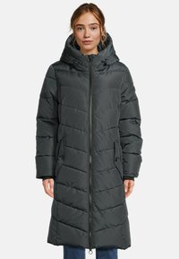 Femme portant un long manteau matelassé noir avec capuche et fermeture éclair à l'avant, se tenant devant un fond blanc uni.