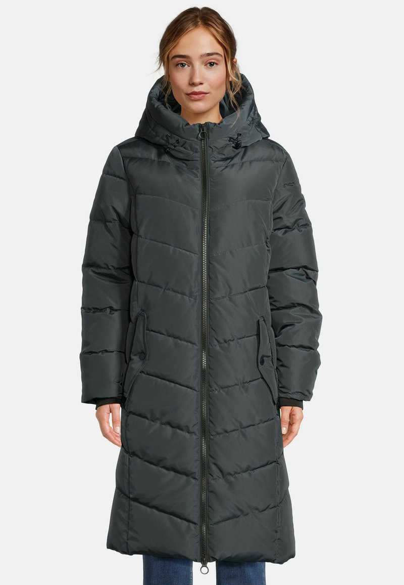 Femme portant un long manteau matelassé noir avec capuche et fermeture éclair à l'avant, se tenant devant un fond blanc uni.