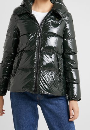 Femme portant une doudoune noire brillante avec fermeture éclair à l'avant et poches, associée à un jean bleu et une chemise blanche en dessous.