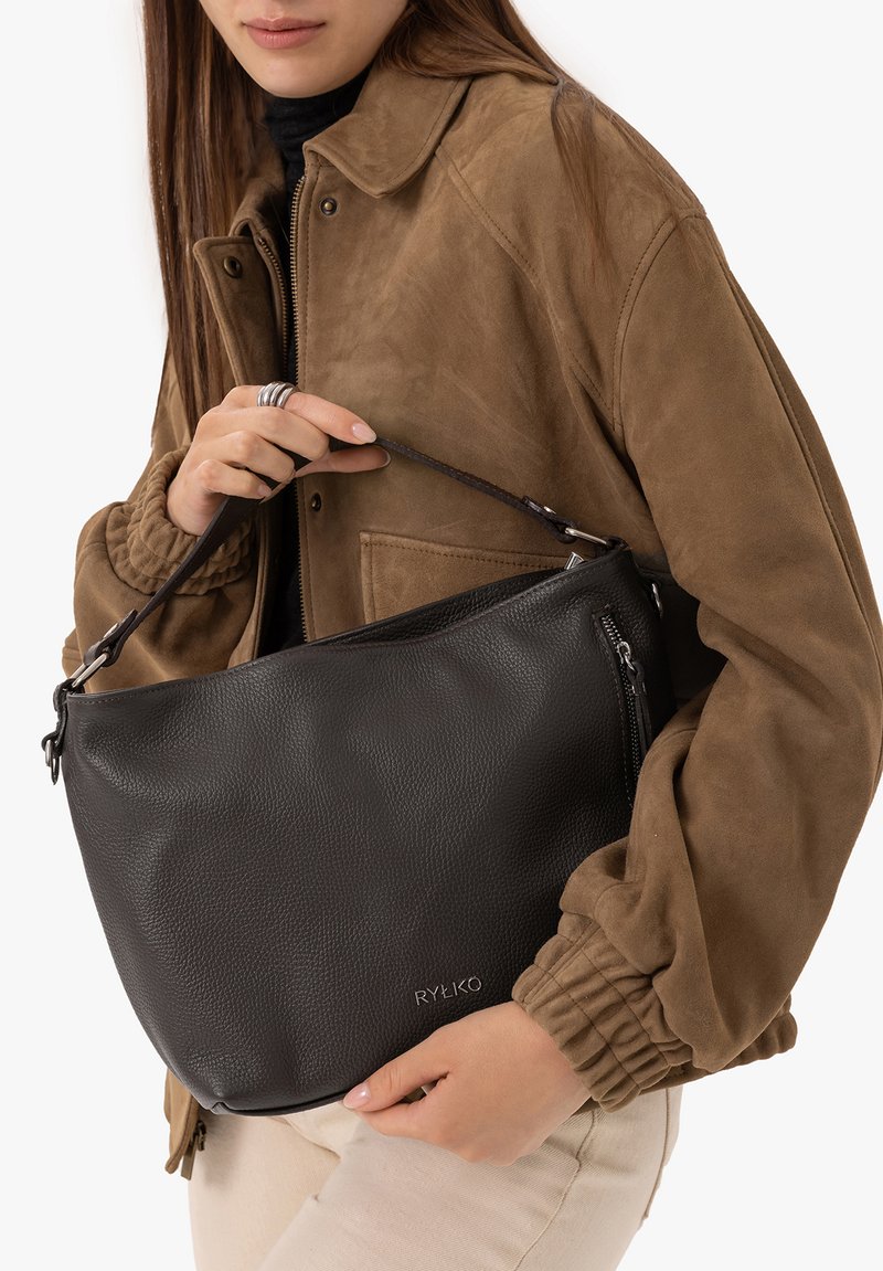 Femme en veste marron tenant un sac à bandoulière en cuir noir avec poche zippée et logo Ryłko.