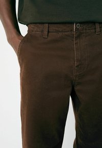 Close-up of brown pants with button and belt loops, worn by person with left hand in pocket, against plain light background.