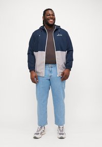 Rompevientos color bloqueado en navy y gris, con cremallera frontal, capucha con cordón y bolsillos laterales, combinado con jeans de color azul claro y zapatillas grises.