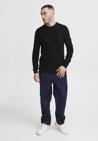 Suéter negro de punto con cuello redondo y mangas largas, combinado con pantalones de mezclilla azul oscuro y zapatillas blancas con detalles amarillos.