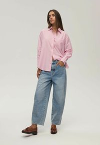 Różowa w paski koszula z guzikami, z jedną kieszonką na piersi, w połączeniu z luźnymi, jasnoniebieskimi dżinsami oraz brązowymi mokasynami.