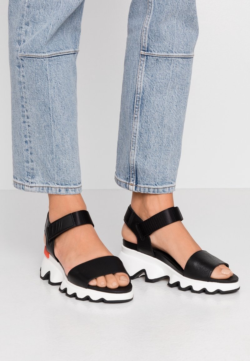 Sandales noires à bout ouvert avec des semelles épaisses blanches en zigzag et des brides à la cheville, portées avec un jean court bleu clair sur pieds nus.