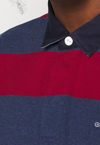 Blau-rot gestreiftes Poloshirt mit dunklem Kragen, Knopfleiste und weichem Material. Logo sichtbar auf der unteren rechten Seite.