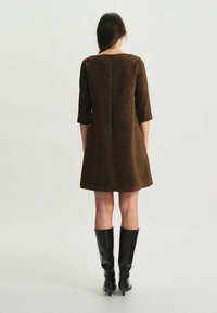 Robe droite en tissu texturé marron avec des manches trois-quarts. Présente une couture arrière droite et est portée avec des bottes noires jusqu'aux genoux.