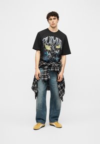 Νέος άνδρας που στέκεται φορώντας μαύρο γραφικό t-shirt Puma, μπλε τζιν, κίτρινα αθλητικά παπούτσια και ένα ασπρόμαυρο καρό πουκάμισο δεμένο γύρω από τη μέση του.