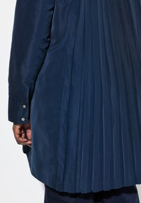 Cappotto plissettato blu navy con una texture del tessuto liscia, dotato di dettagli con bottoni a scatto sui polsini e una silhouette rilassata e fluida.