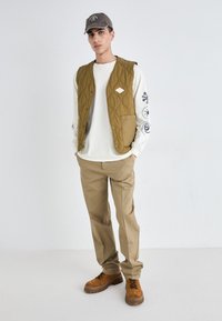 Quilted olive vest over white long-sleeve shirt, beige pants, and brown hiking boots. The vest has large pockets and a front snap closure.
