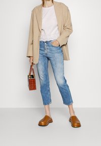 Beige linnblazer över en vit topp med ögla detaljer, ljusblå cropped jeans med knappslut, och bruna träskor. Liten brun handväska inkluderad.