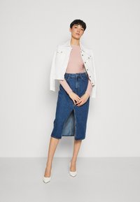 Weißer Jeansjacke über einem rosa gerippten Oberteil, kombiniert mit einem knielangen blauen Jeansrock mit Schlitz vorne, ergänzt durch weiße Absätze.