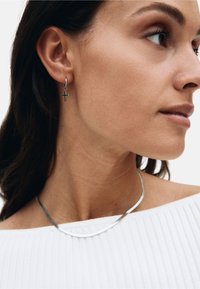 Silver hoops med en liten korsberlock; elegant silversmycke; modell bär en vit ribbad off-shoulder topp.