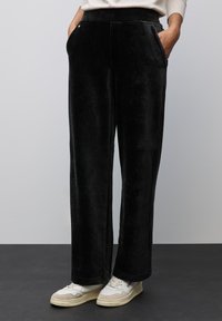 Pantalons larges noirs en matière douce et veloutée, avec des poches avant et une taille texturée, associés à des baskets blanches et beige.