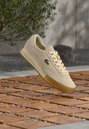 Beige sneaker with textured upper, light laces, and a gum sole. Features a logo on the side and a streamlined silhouette.