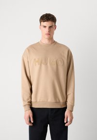 Sweatshirt beige avec col ras du cou et poignets côtelés, arborant un texte en relief "HUGO" sur le devant. Assorti avec un jean noir.