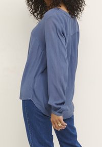 Hellblaue Bluse mit langen Ärmeln aus strukturiertem Stoff, lässiger Schnitt und seitlichen Schlitzen. Getragen mit blauen Jeans, die eine entspannte Silhouette zeigen.