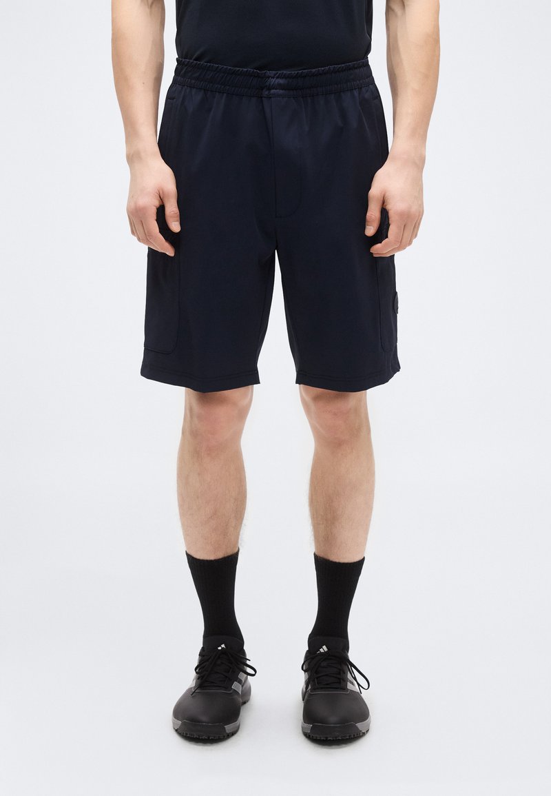 Σκούρο μπλε αθλητικό σορτς με ελαστική μέση, λείο ύφασμα, πλαϊνές τσέπες και ένα λογότυπο στην αριστερή πλευρά. Μαύρα αθλητικά παπούτσια και κάλτσες.